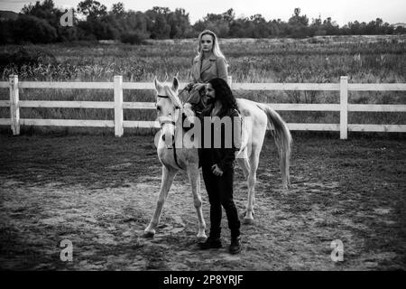 Ein Paar, das mit einem Pferd auf dem Land spaziert. Ein gutaussehender bärtiger Mann, der ein Pferd für einen Zauber fährt und seine Freundin ansieht. Frau reitet graue araberin Stockfoto