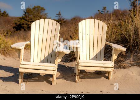 Zwei weiße Adirondack-Stühle auf einem Sandstrand in New England, Cape Code, mit Vegetation im Hintergrund Stockfoto