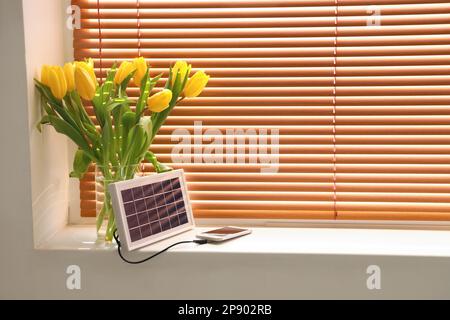 Tragbares Solarpaneel-Mobiltelefon und Vase mit Tulpen auf Fensterbank im Zimmer Stockfoto