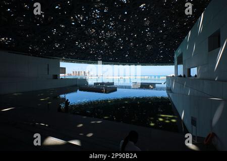Der Blick unter der gewobenen Metallkuppel auf den großen reflektierenden Pool und die Entwicklung, die Bauarbeiten dahinter. Im Kunstmuseum des französischen Louvre in Abu DH Stockfoto