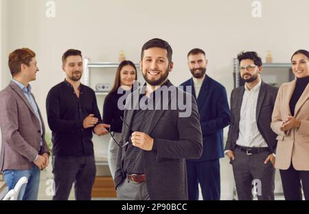 Glückliche Mitarbeiter begrüßen einen aufregenden, gut aussehenden brünetten Mann in schwarzem Anzug und begrüßen ein neues Teammitglied bei der Arbeit. Stockfoto