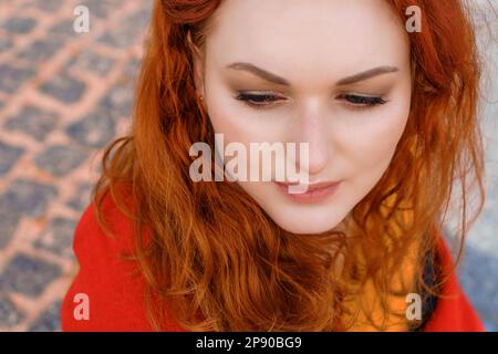 Nahaufnahme Porträt junge weiße Frau mit rotem lockigem Haar in einem roten Mantel im Wind entwickelt Locken und genießt die Natur Stockfoto