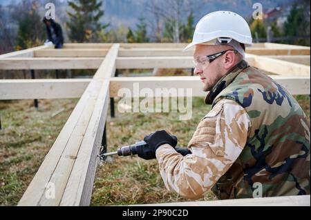 Ein Mann, der ein Holzrahmenhaus auf einem Pfahlgrundstück baut. Männlicher Arbeiter bohrt mit einem Elektrobohrer ein Loch in die Holzrahmung des zukünftigen Hauses. Baumeister trägt weißen Helm und Schutzbrille. Zimmereikonzept. Stockfoto