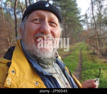 Freundlicher älterer Mann mit Bart und Dockers Kappe, der in die Kamera schaut. Er nahm seine Brille ab. Er wacht mit gelbem Regenmantel im Wald auf. Stockfoto