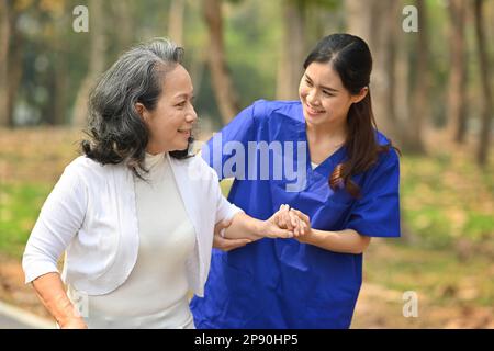 Asiatische Krankenschwester in Uniform, die Hand einer älteren Frau hält und während des Spaziergangs im Rehabilitationszentrum ein angenehmes Gespräch führt Stockfoto