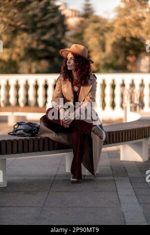 Outdoor-Modeporträt einer jungen eleganten, modischen braunen Frau, Model mit stilvollem Hut, Würfelchen und leichter Regenjacke, die bei Sonnenuntergang in Europa posiert Stockfoto