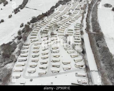 Ripon, North Yorkshire, Großbritannien. März 2023. Aus der Vogelperspektive zeigt einen schneebedeckten Campingplatz mit Wohnwagen und Stellplätzen neben angrenzenden Stadtgebieten von Ripon. Schneedecken decken Dächer, Straßen, Felder und Gebäude in der Stadt. Das Foto wurde aus einer erhöhten Position an einem klaren Wintertag nach nächtlichem Schneefall aufgenommen. Das Bild zeigt den Kontrast zwischen dem weißen Campingplatz und den teilweise schneebedeckten Wohn- und Geschäftszonen dieser historischen Marktstadt in North Yorkshire. Im Rahmen sind keine Personen sichtbar. Stockfoto