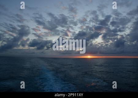 Sonnenuntergang am Atlantik mit verstreuter Wolke am blauen Himmel und einem Kreuzfahrtschiff, das von Mitte nach links verläuft. Sonne in einer kleinen Lücke zwischen Wolke und Horizont. Stockfoto