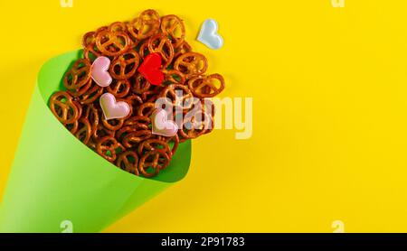 Mini-Brezeln und kleine bunte Herzen in hellgrünem Papierkegel liegen auf gelbem Hintergrund. Nahaufnahme-Banner mit Kopierbereich. Stockfoto