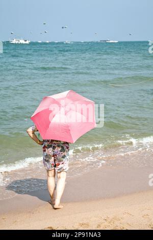 Ältere Frau in kurzen Hosen mit pinkfarbenem Sonnenschirm am Strand, Sonnenbaden, Blick auf den Horizont Stockfoto