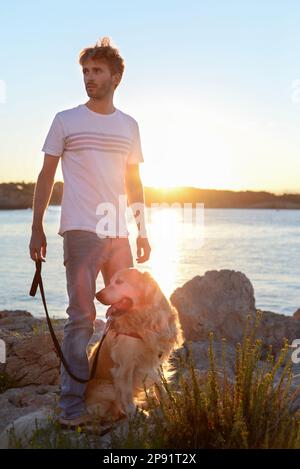 Hübscher Erwachsener genießt den Sonnenuntergang mit seinem Hund zusammen am Meer. Golden Retriever und sein Mensch haben eine große Freundschaft. Stockfoto