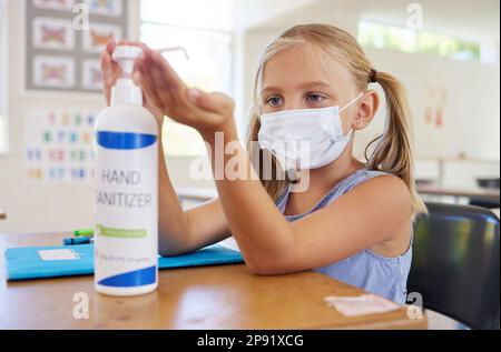 Ein kleiner Student desinfiziert die Hand, um sich vor Covid zu schützen, trägt eine Maske und folgt einer neuen Hygienesicherheitsroutine. Kindergarten oder Vorschule weiblich Stockfoto