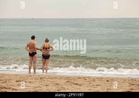 Paar mittleren Alters am Meer Wasser halten sich an den Händen. Kaukasische Mann und Frau am Strand von hinten. Idyllischer Seenlandschaft mit Menschen Stockfoto