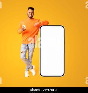 Ein hübscher indianer, der neben einem riesigen Smartphone steht, mit leerem Bildschirm, lächelt und hält das Telefon isoliert auf gelbem Hintergrund, Modell, Kopierraum Stockfoto