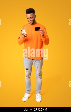 Ein fröhlicher, gutaussehender Kerl, der für den lang erwarteten Kauf auf dem Handy bezahlt, mit Kreditkarte und auf den Bildschirm schaut. Ein glücklicher Mann aus dem Nahen Osten bestellt online Essen, kauft ein, volle Länge Stockfoto