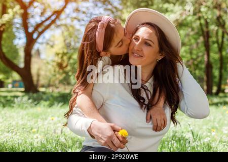 Das Mädchen küsst die Mutter auf der Wange und umarmt sie, während sie im Frühlingspark auf dem Gras sitzt und Blumen schenkt. Entspannung für die ganze Familie im Freien. Muttertag. Schließen Stockfoto