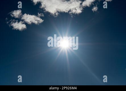 Die Sonne scheint hell in Sternform am hellblauen Himmel mit ein paar weißen Wolken als horizontalem Hintergrund Stockfoto