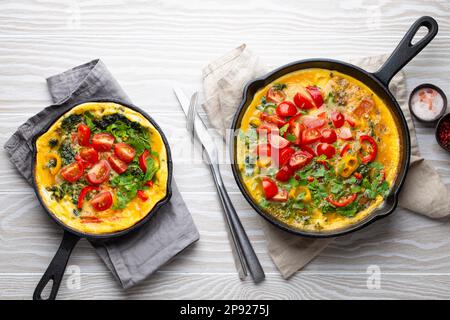 Gesunde Frittata in zwei gusseisernen Pfannen mit gebratenen Eiern und Gemüse der Saison auf rustikalem weißem Holzhintergrund. Italienisches Omelette mit Stockfoto