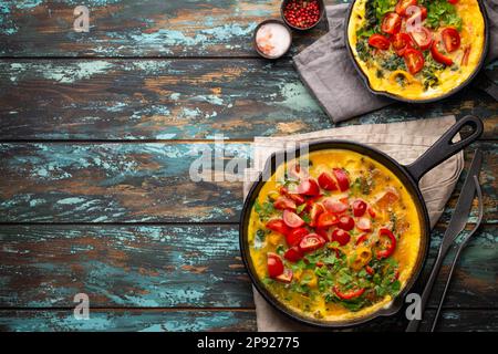 Gesunde Frittata in zwei gusseisernen Pfannen mit gebratenen Eiern und Gemüse der Saison auf rustikalem Holzhintergrund. Italienisches Omelette mit Bio-Omelette Stockfoto