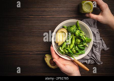 Schüssel mit gesundem Salat in weiblichen Händen von oben und grüner Detox Smoothie in einer Flasche auf Holzhintergrund, Draufsicht. Eine Frau isst leckeres veganes Essen Stockfoto