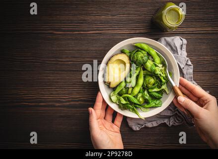 Schüssel mit gesundem Salat in weiblichen Händen von oben und grüner Detox Smoothie in einer Flasche auf Holzhintergrund, Draufsicht. Eine Frau isst leckeres veganes Essen Stockfoto