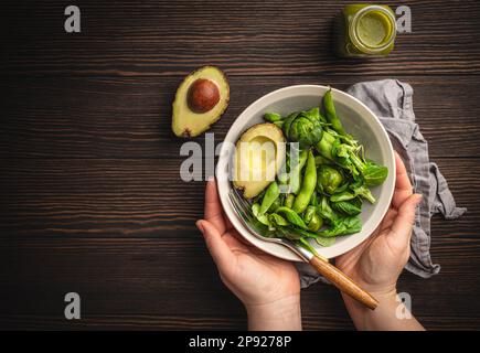 Schüssel mit gesundem Salat in weiblichen Händen von oben und grüner Detox Smoothie in einer Flasche auf Holzhintergrund, Draufsicht. Eine Frau isst leckeres veganes Essen Stockfoto