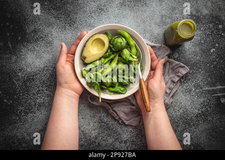 Schüssel mit gesundem Salat in weiblichen Händen von oben und grüner Detox Smoothie in einer Flasche auf grauem Steinhintergrund, Draufsicht. Eine Frau, die leckere Veganer isst Stockfoto