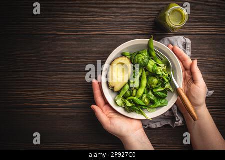 Schüssel mit gesundem Salat in weiblichen Händen von oben und grüner Detox Smoothie in einer Flasche auf Holzhintergrund, Draufsicht. Eine Frau isst leckeres veganes Essen Stockfoto