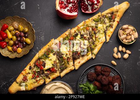 Traditionelle türkische Pizza, frisch gebacken mit Rinderhack und Gemüse, serviert auf schwarzem Steinhintergrund mit verschiedenen Meze aus dem Nahen Osten Stockfoto