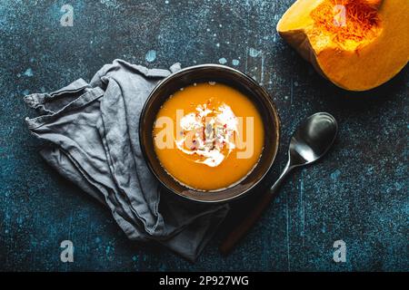 Hausgemachte Kürbiscremesuppe in brauner Keramikschüssel auf dunklem Steinhintergrund mit Löffel, dekoriert mit geschnittenem frischen Kürbis, Draufsicht. Traditionell Stockfoto