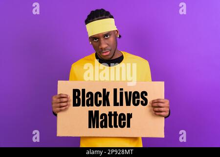 Schwarzer ethnischer Mann in gelber Kleidung auf lila Hintergrund, zeigt mit einem Schild, schwarze Leben sind wichtig Stockfoto
