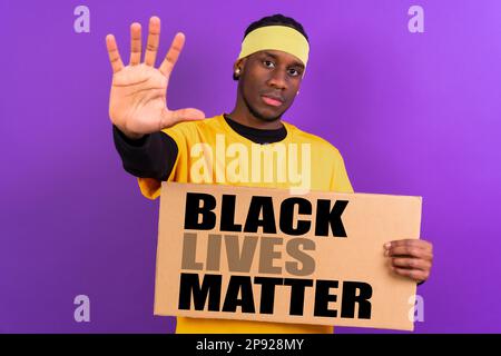 Schwarzer ethnischer Mann in gelber Kleidung auf lila Hintergrund, zeigt mit einem Schild, schwarze Leben sind wichtig, hält mit seiner Hand an Stockfoto