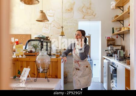 Junge Barista, die hinter der Bar im Café steht und über ein Smartphone spricht, Konzept für Inhaber eines kleinen Unternehmens, authentisches Design. Stockfoto