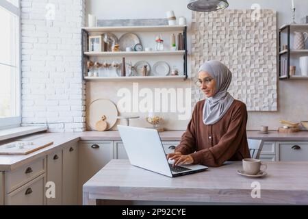 Online-Arbeit von zu Hause aus. Eine junge muslimische Frau in einem Hidschab sitzt zu Hause in der Küche am Tisch und arbeitet und lernt von zu Hause aus mit einem Laptop. Stockfoto