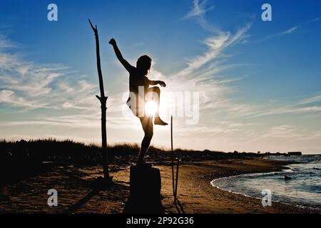 Der Junge übt Meditation und Yoga an einem sonnigen Sommerstrand. Lotus-Position in Silhouettenstellung. Stockfoto