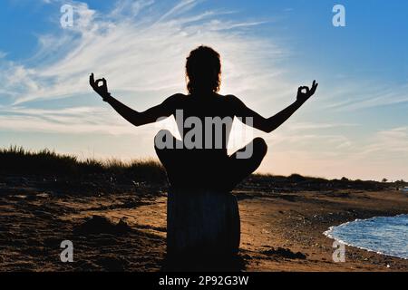 Der Junge übt Meditation und Yoga an einem sonnigen Sommerstrand. Lotus-Position in Silhouettenstellung. Stockfoto