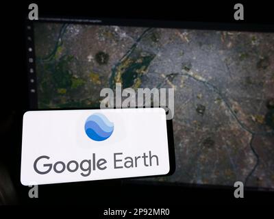 Person, die ein Mobiltelefon mit dem Logo der Satellitenbildsoftware Google Earth auf dem Bildschirm vor der Webseite hält. Konzentrieren Sie sich auf das Display des Telefons. Stockfoto