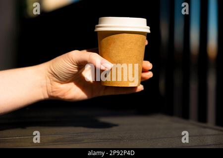 Die Hand einer Frau hält eine Papptasse Kaffee mit weißem Deckel. Sommer, sonniger Tag. Stockfoto