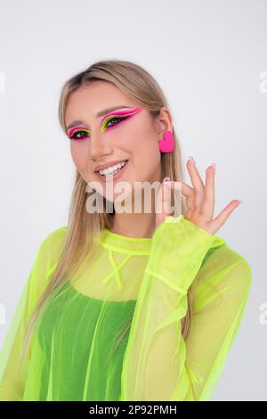 Es ist alles gut. Eine junge Blondine mit leuchtendem Neon-Make-up lächelt und zeigt mit ihren Fingern, dass alles in der Kamera gut läuft. Stockfoto