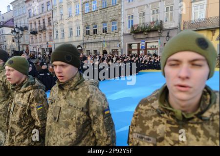 Lemberg, Ukraine. 10. März 2023. Militärpersonal, Kadetten und Polizisten singen die Nationalhymne, während sie vor dem Rathaus Ratusha eine riesige ukrainische Flagge halten, um den Jahrestag der ersten Aufführung der Nationalhymne vor 158 Jahren zu feiern. (Foto: Mykola Tys/SOPA Images/Sipa USA) Guthaben: SIPA USA/Alamy Live News Stockfoto