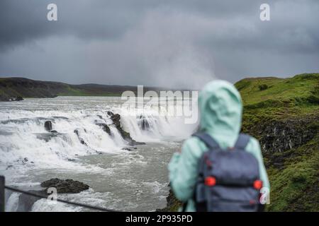 Frau mit Rucksack und grüner Jacke, die vor dem mächtigen Wasserfall Gullfoss in Island steht. Touristenattraktion, die der Golden Circle Route von Island folgt. Stockfoto