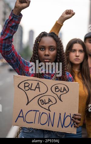 Eine junge afrikanische Frau nimmt an einem protestmarsch Teil und hält ein Schild mit der Aufschrift „Bla Bla...Act Now!“. Sie hebt ihre Faust in die Luft, als Zeichen von P. Stockfoto