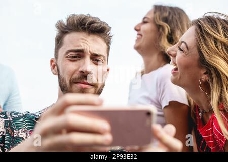 Ein junger Mann mit Bart sieht auf sein Handy mit einem gestörten Ausdruck, während zwei Mädchen im Hintergrund lachen. Ein Moment von Freunden, die Smartphones benutzen Stockfoto