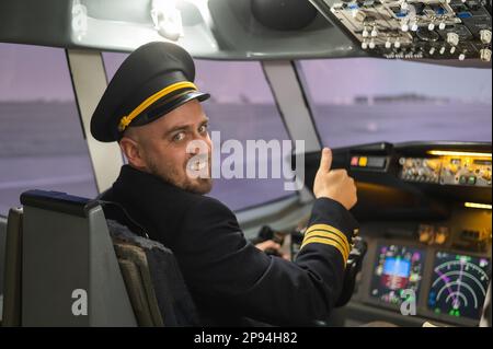 Kaukasischer bärtiger Mann, der beim Fahren eines Flugsimulators lächelt. Der Pilot im Cockpit zeigt Daumen nach oben. Stockfoto