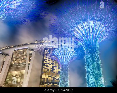 SINGAPUR - 14. JULI 2022: Elektrisches blaues Licht von supertrees in the Gardens am Bay Park in Singapur erhellt den Nachthimmel mit dem M Stockfoto