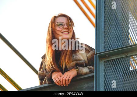 Eine junge Frau mit rötlichen Haaren und Brille in karierter Hemdjacke neigt sich über das überdachte Geländer und blickt in die Ferne, von unten gesehen Stockfoto