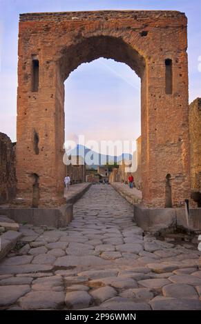 Der Caligula-Bogen - Arco di Caligola - in der Via Di Mercurio, Pompeji. Kopfsteinpflasterstraßen und Vesuv in der Ferne. Pompeji wurde nach dem katastrophalen Ausbruch des Vesuv im Jahr 79 n. Chr. unter Meter von Asche und Bimsstein begraben Die erhaltene Stätte bietet ausgegrabene Ruinen von Straßen und Häusern, die Besucher frei erkunden können. Stockfoto