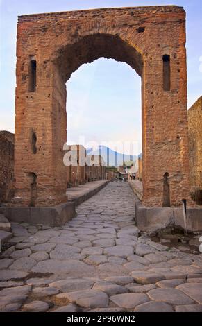 Der Caligula-Bogen - Arco di Caligola - in der Via Di Mercurio, Pompeji. Kopfsteinpflasterstraßen und Vesuv in der Ferne. Pompeji wurde nach dem katastrophalen Ausbruch des Vesuv im Jahr 79 n. Chr. unter Meter von Asche und Bimsstein begraben Die erhaltene Stätte bietet ausgegrabene Ruinen von Straßen und Häusern, die Besucher frei erkunden können. Stockfoto