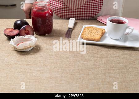 Die Person bereitet das Frühstück mit Marmeladenglas, frischen Pflaumen, Toast und Teetasse auf dem rustikalen Tisch zu. Stockfoto