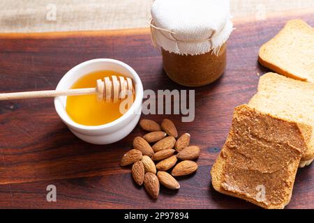 Rustikale Frühstücksszene: Honig in der Schüssel, Mandelbutter, Mandeln, Toast auf Holz. Köstlich und gesund. Stockfoto